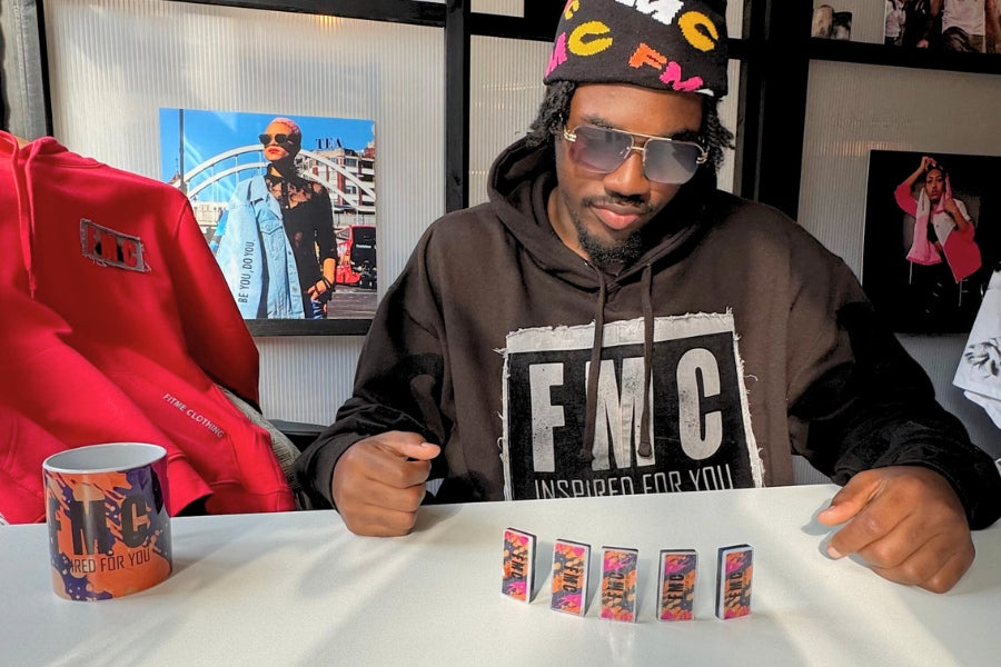 A male wearing a hoodie with 'FMC' branded hoodie, sitting at a table playing dominos at a FitMe Clothing pop up event.